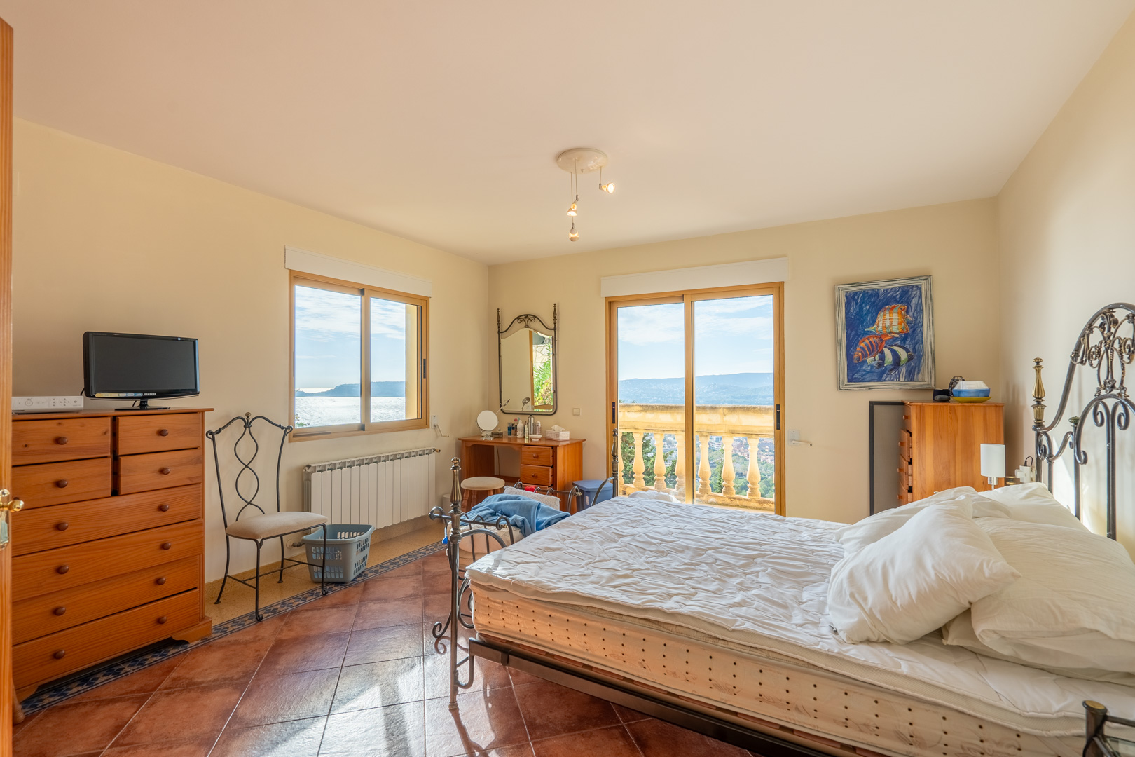 Chambre avec sol en terre cuite, lit en fer forgé et accès à un balcon avec balustrade offrant une vue dégagée sur la baie et la mer Méditerranée.
