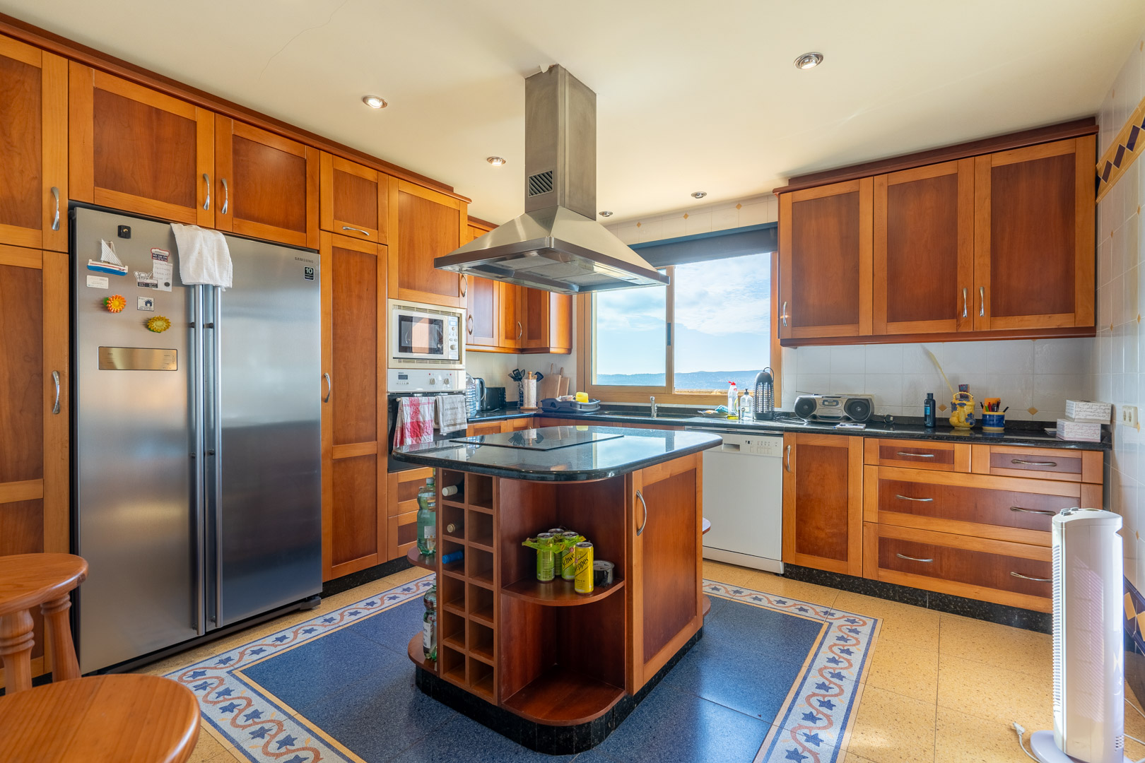 Cuisine traditionnelle avec meubles en bois, îlot central avec plan de travail en granit noir et hotte en inox. Elle offre des appareils intégrés et une vue sur la mer par la fenêtre.