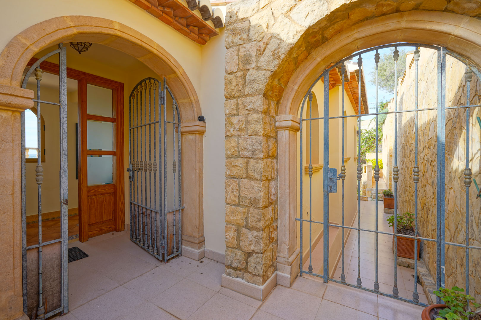 Entrée avec arches en pierre de Tosca et grilles en fer forgé menant à la porte principale en bois et à un passage latéral extérieur.