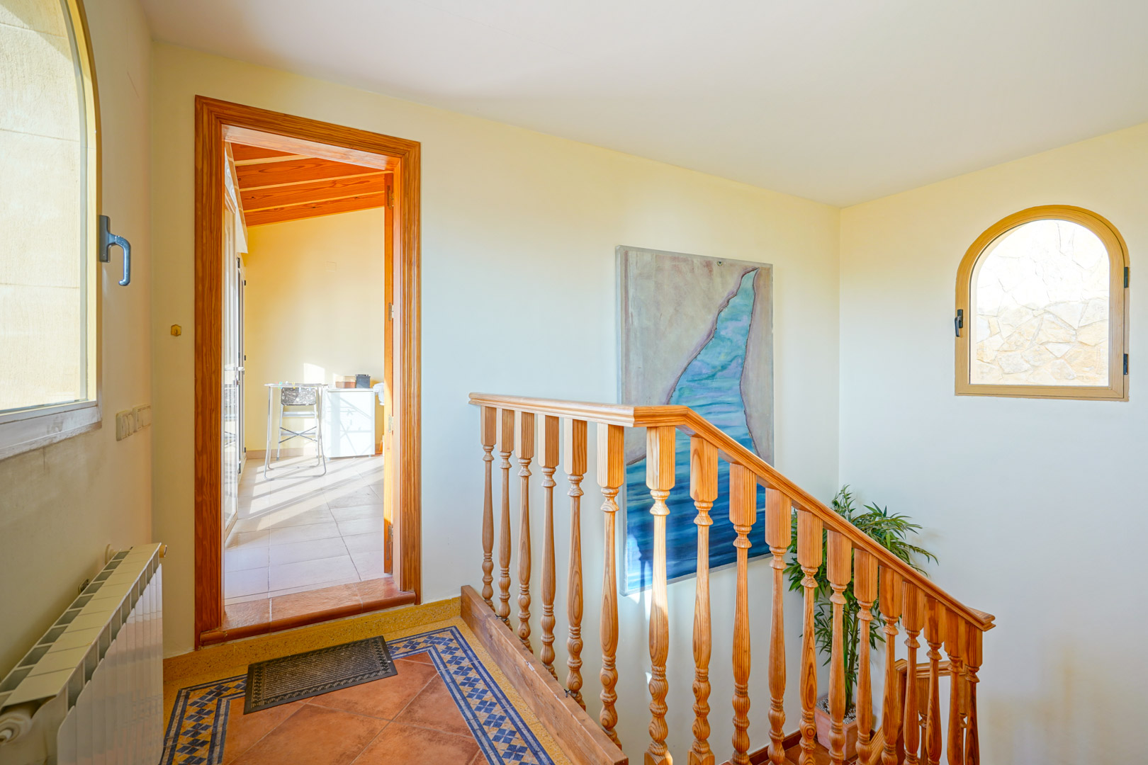 Palier avec escalier en bois, sol en carrelage décoratif et fenêtre en arche. L'espace s'ouvre sur une pièce lumineuse dotée d'un plafond mansardé en bois.