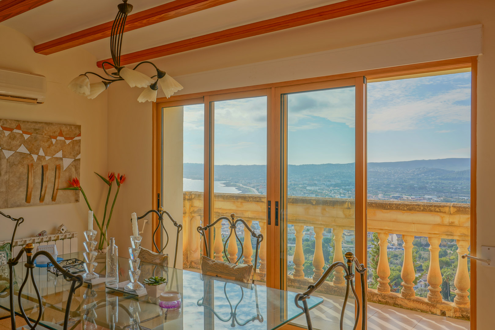 Salle à manger avec table en verre et poutres en bois, dotée de grandes baies vitrées s'ouvrant sur une vue panoramique de la baie de Jávea.