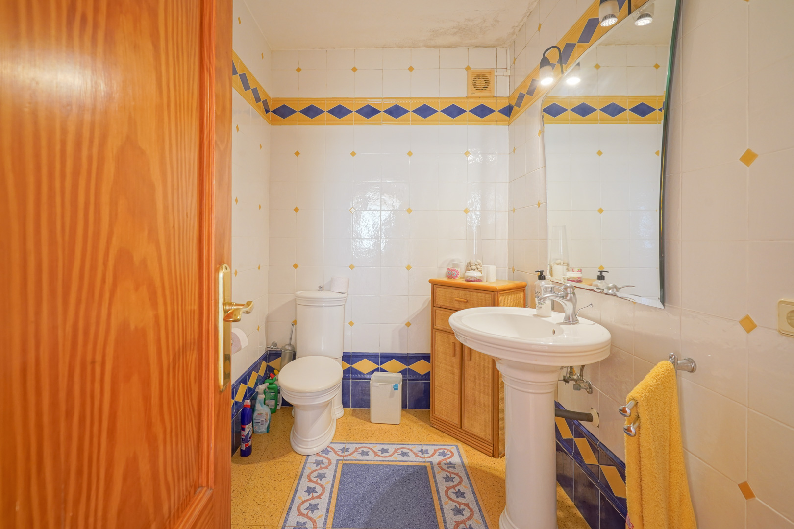 Salle de bain traditionnelle avec carrelage blanc aux bordures bleues et jaunes, lavabo sur colonne et petit meuble de rangement en bois.