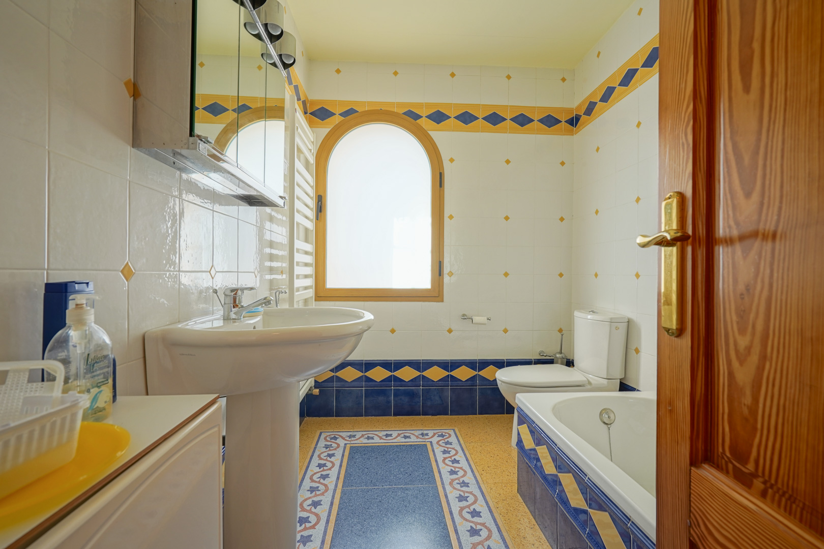 Salle de bains avec carrelage décoratif bleu et jaune, fenêtre cintrée en bois et agencement comprenant une baignoire, un lavabo sur colonne et un radiateur sèche-serviettes.