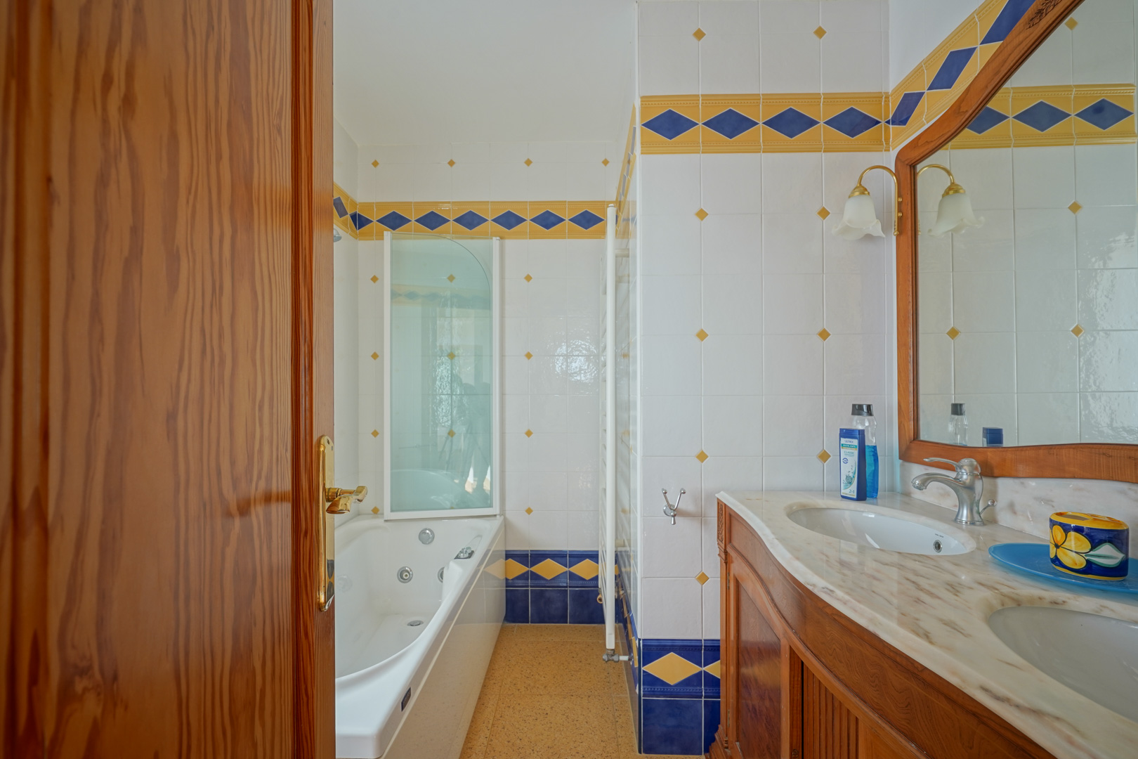 Salle de bains avec double vasque en marbre, meuble en bois et baignoire d'hydromassage, décorée de carreaux aux motifs géométriques bleus et jaunes.
