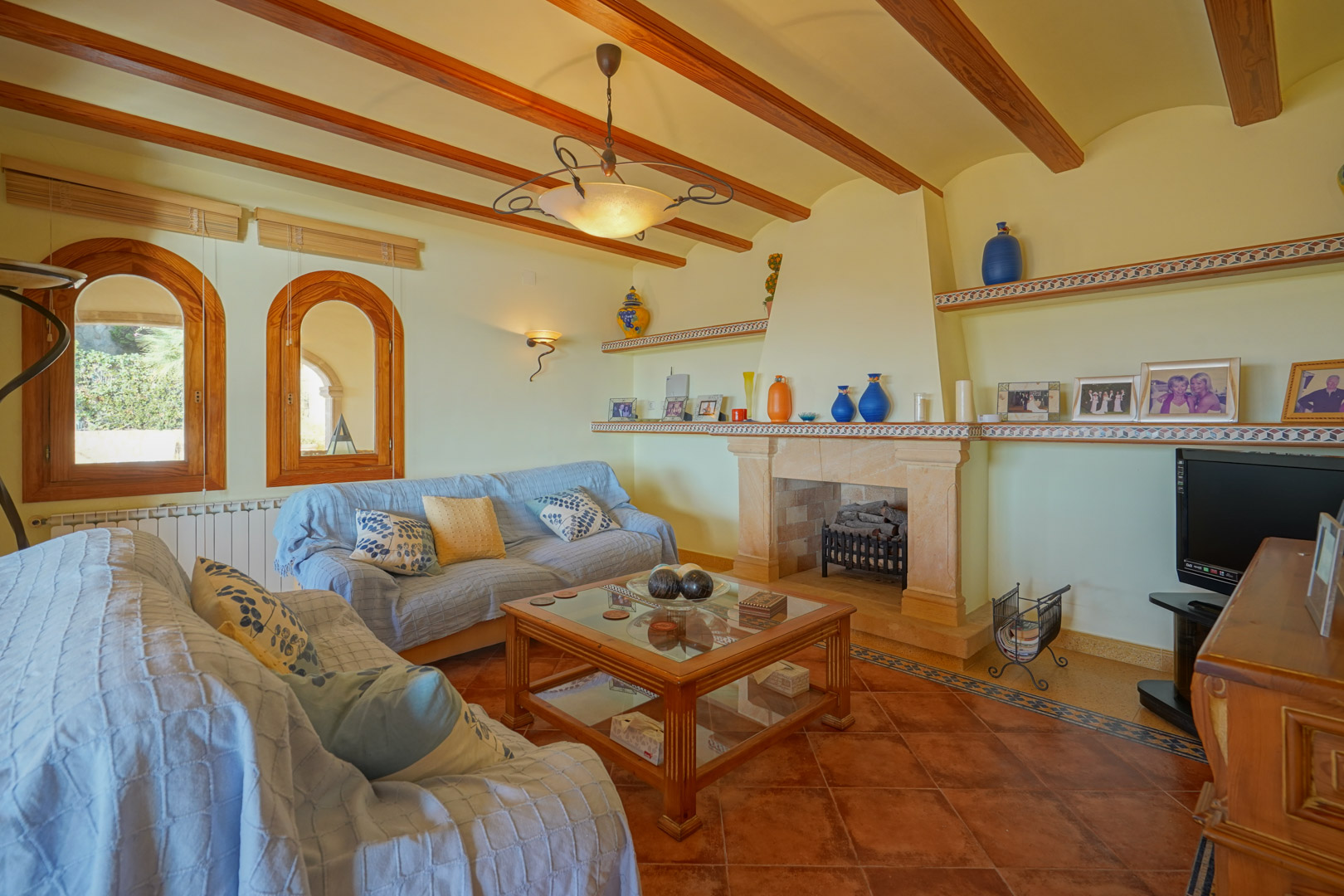 Salon traditionnel avec cheminée en pierre, poutres en bois apparentes et sol en terre cuite. Fenêtres cintrées en bois et étagères intégrées aux détails en céramique.