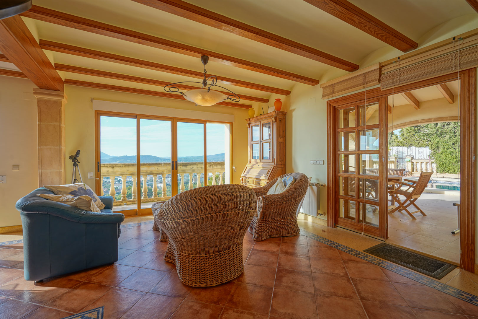 Séjour avec poutres apparentes, sol en terre cuite et fauteuils en osier. Les baies vitrées s'ouvrent sur une terrasse offrant une vue dégagée sur la vallée et les montagnes.