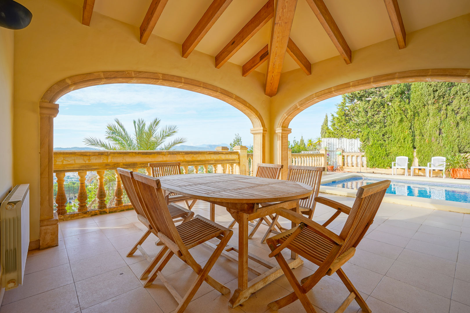 Terrasse couverte avec poutres en bois apparentes et arches en pierre, offrant un espace repas extérieur face à la piscine et aux montagnes.