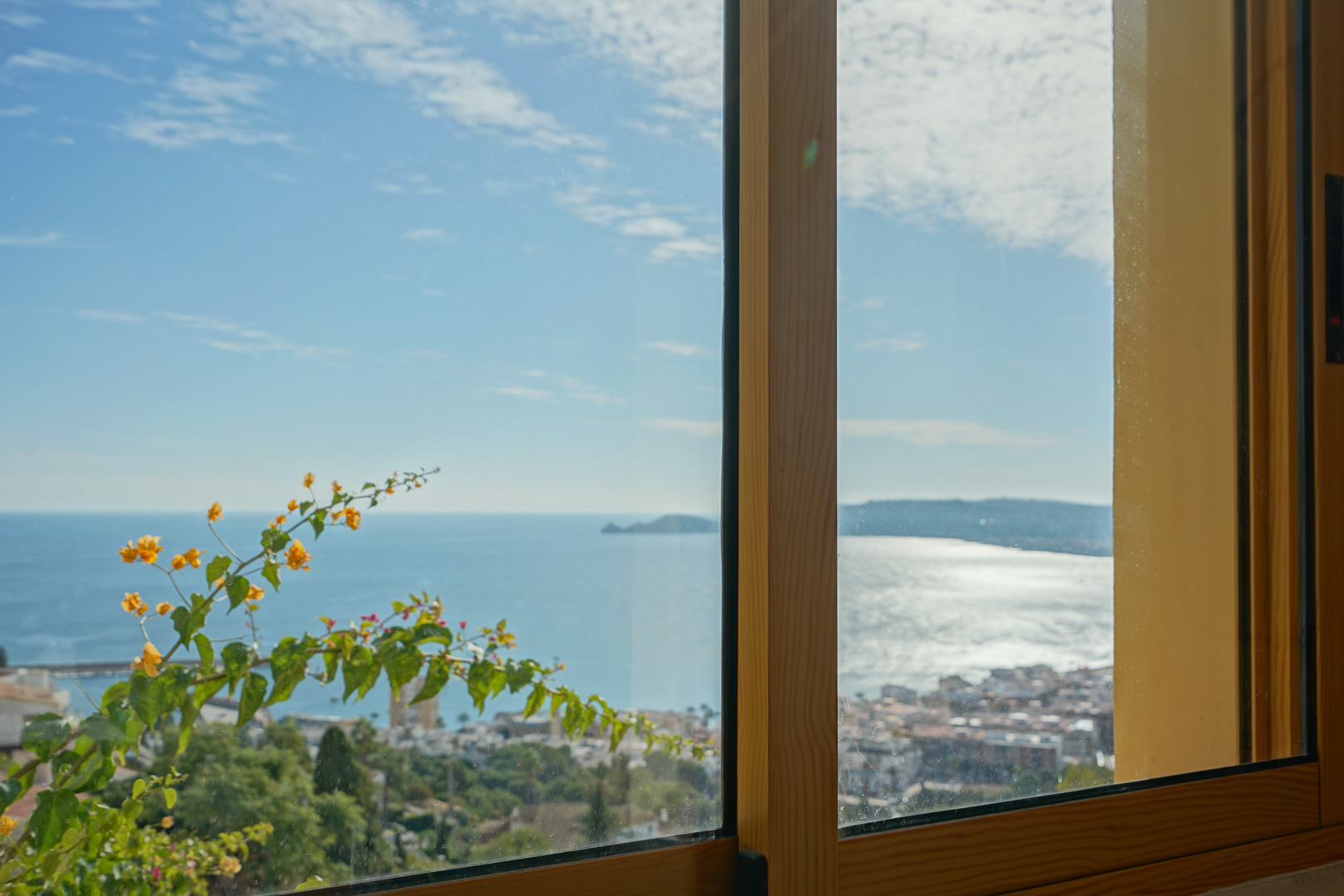 Vue panoramique sur la baie de Jávea et le Cap Negre à travers une fenêtre en bois, agrémentée de bougainvilliers en fleurs.