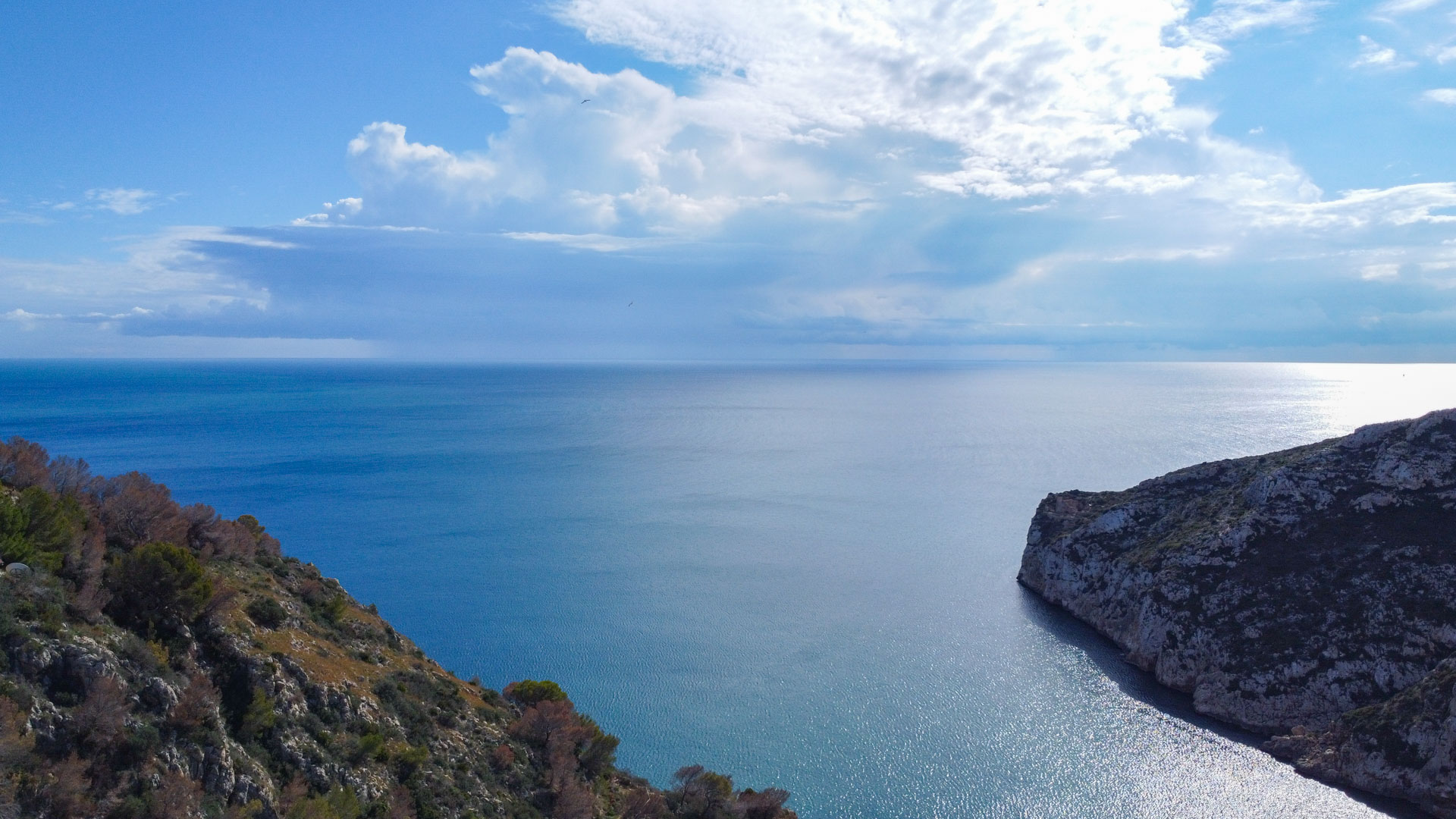 Vue aérienne frontale de la mer Méditerranée et des falaises rocheuses bordant la baie de Granadella sous un ciel partiellement nuageux.