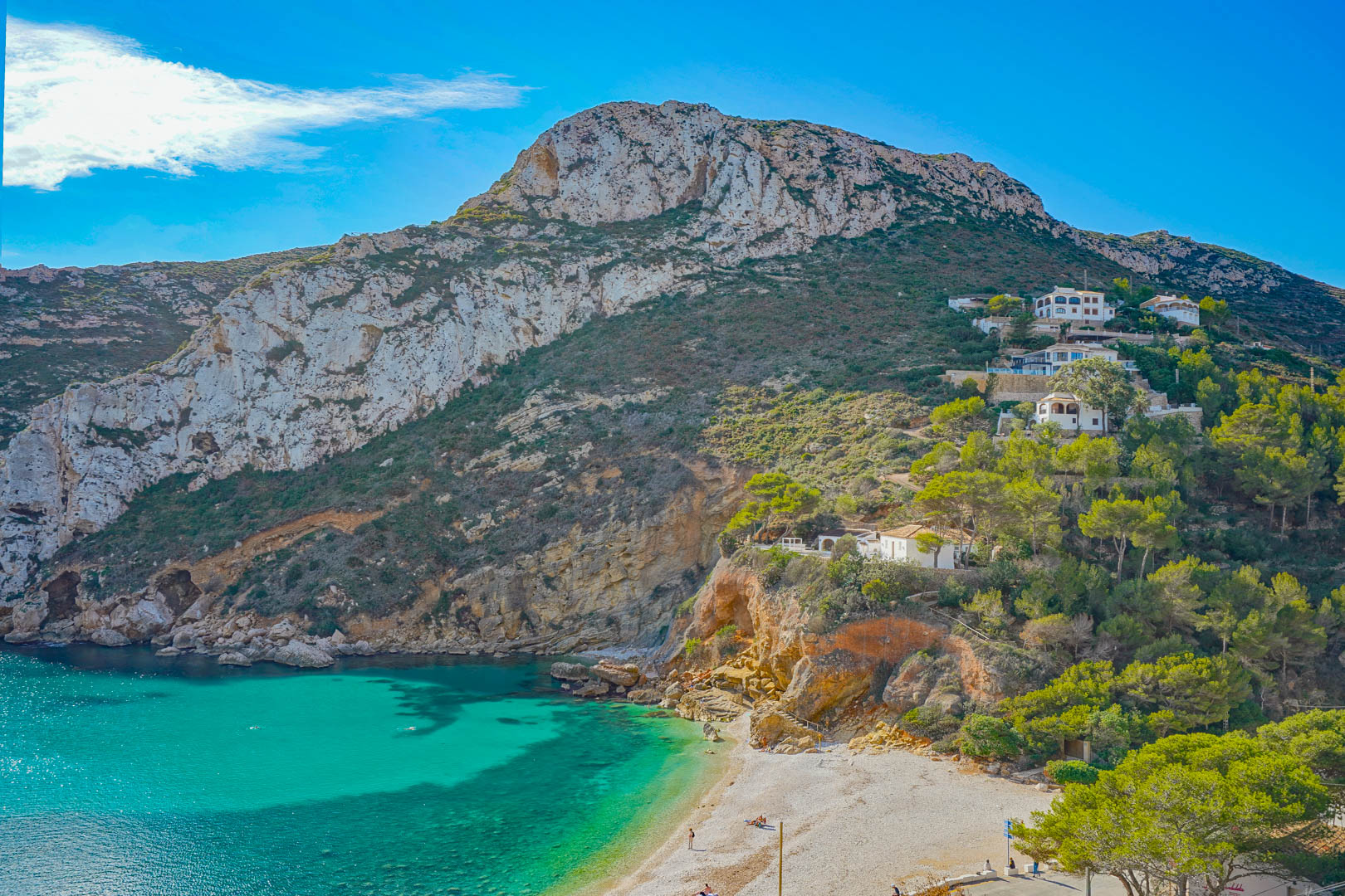 Vue panoramique de la Cala Granadella avec ses eaux turquoise, ses falaises de calcaire et sa végétation méditerranéenne bordant la plage de galets.