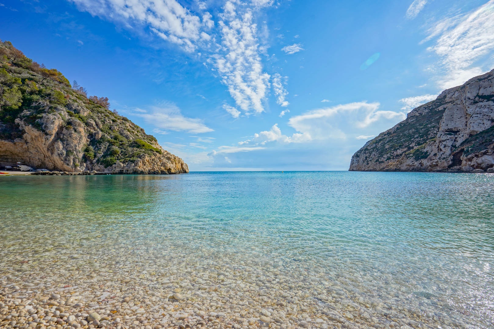 Vue panoramique de la plage de Granadella avec ses eaux turquoise cristallines et son rivage de galets, encadrée par des falaises escarpées et un ciel bleu azur.