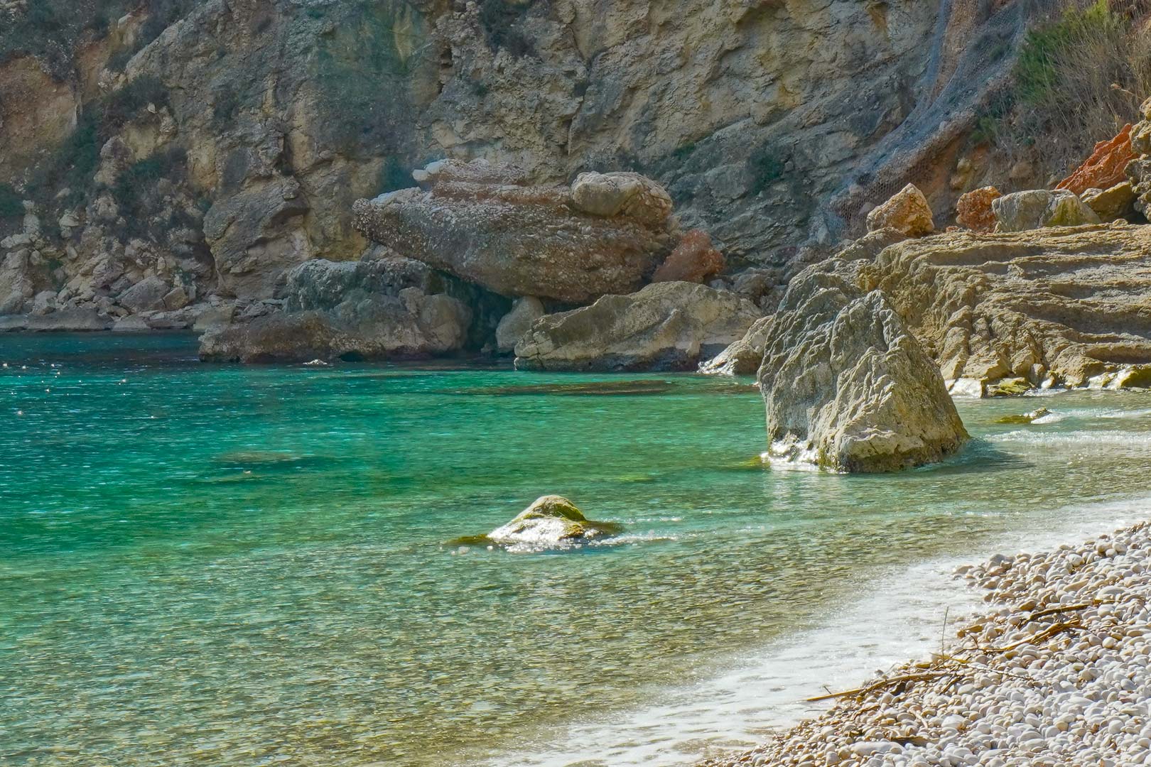Vue sur le rivage de galets de la Cala Granadella, mettant en valeur les eaux turquoise cristallines et les formations rocheuses naturelles environnantes.