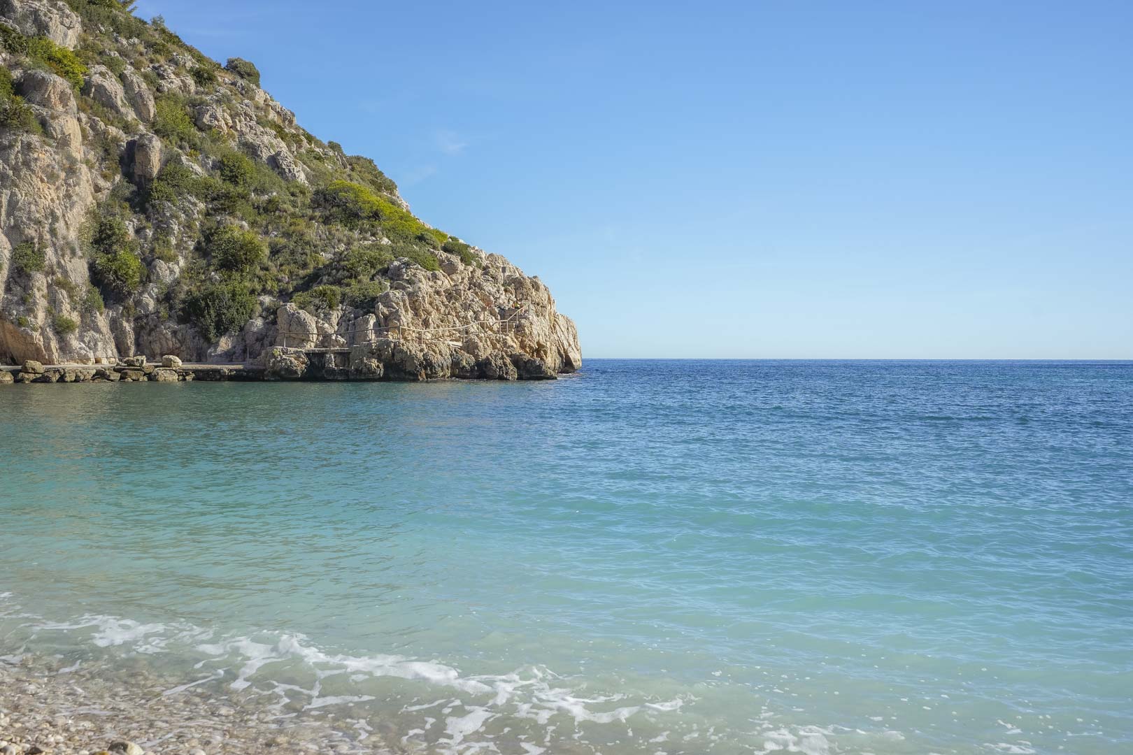 Vue sur les eaux turquoise de la Cala Granadella, bordée par une plage de galets et des falaises escarpées couvertes de végétation méditerranéenne.