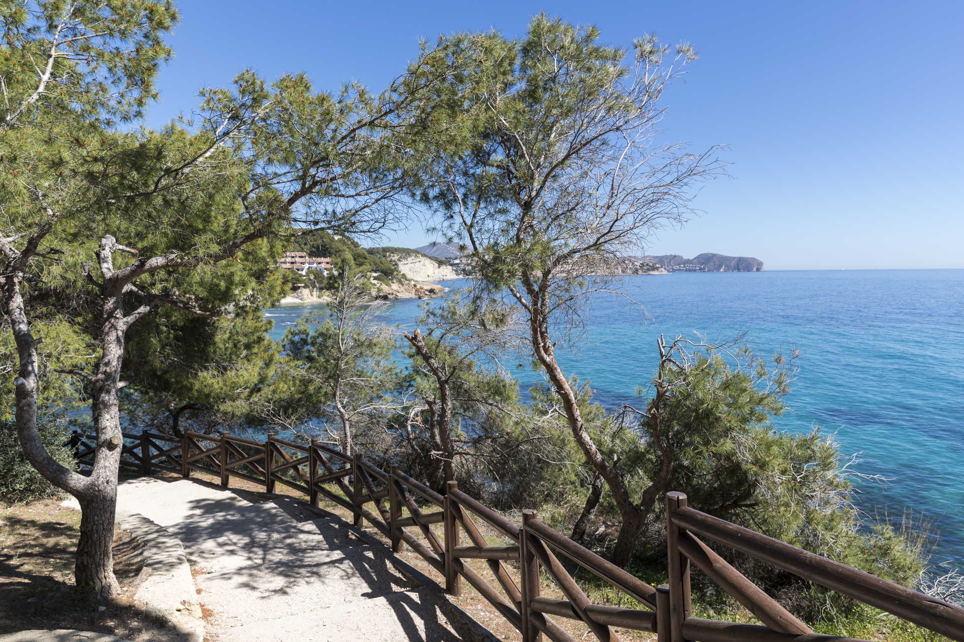 Sentier côtier avec garde-corps en bois et pins parasols, offrant une vue dégagée sur les eaux turquoise de la Méditerranée près de La Fustera.