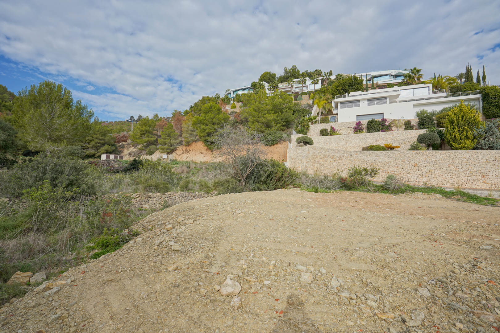 Terrain à bâtir en préparation offrant une vue sur des villas contemporaines blanches et des murs de soutènement en pierre sèche intégrés à la colline boisée.