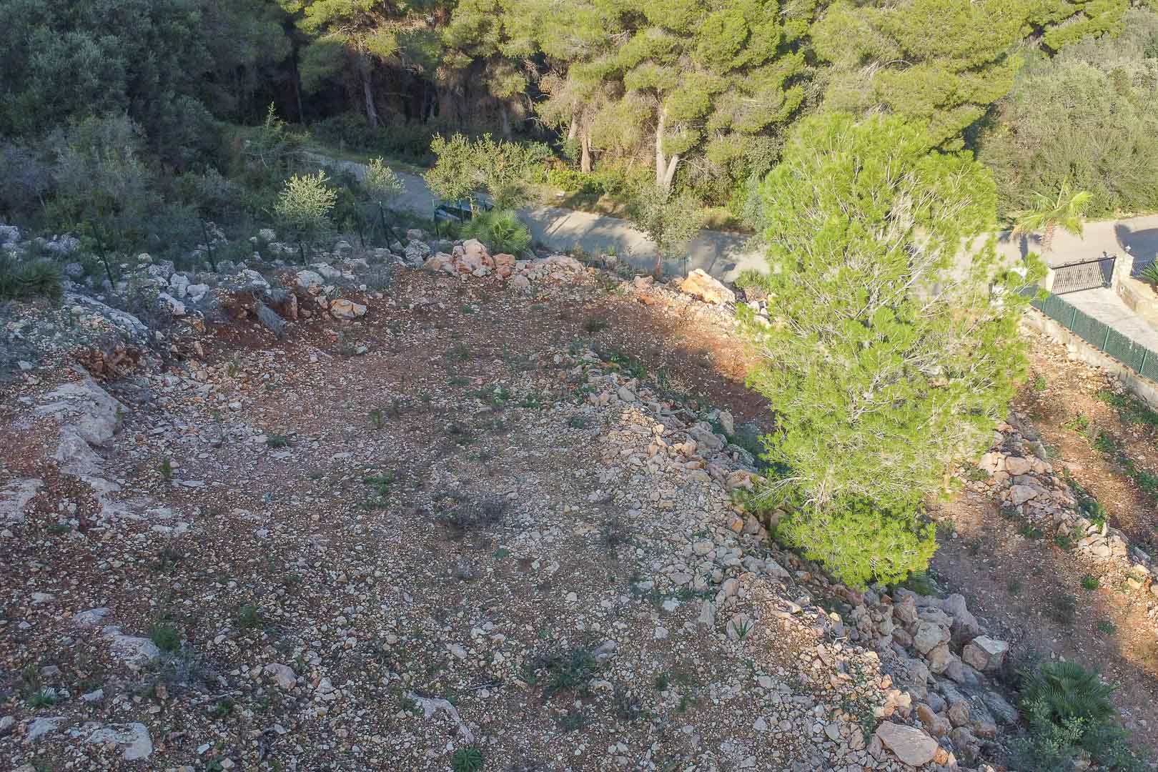 Vue surélevée du terrain à bâtir montrant la topographie naturelle avec terre rouge et rochers, bordée par une forêt de pins méditerranéens.