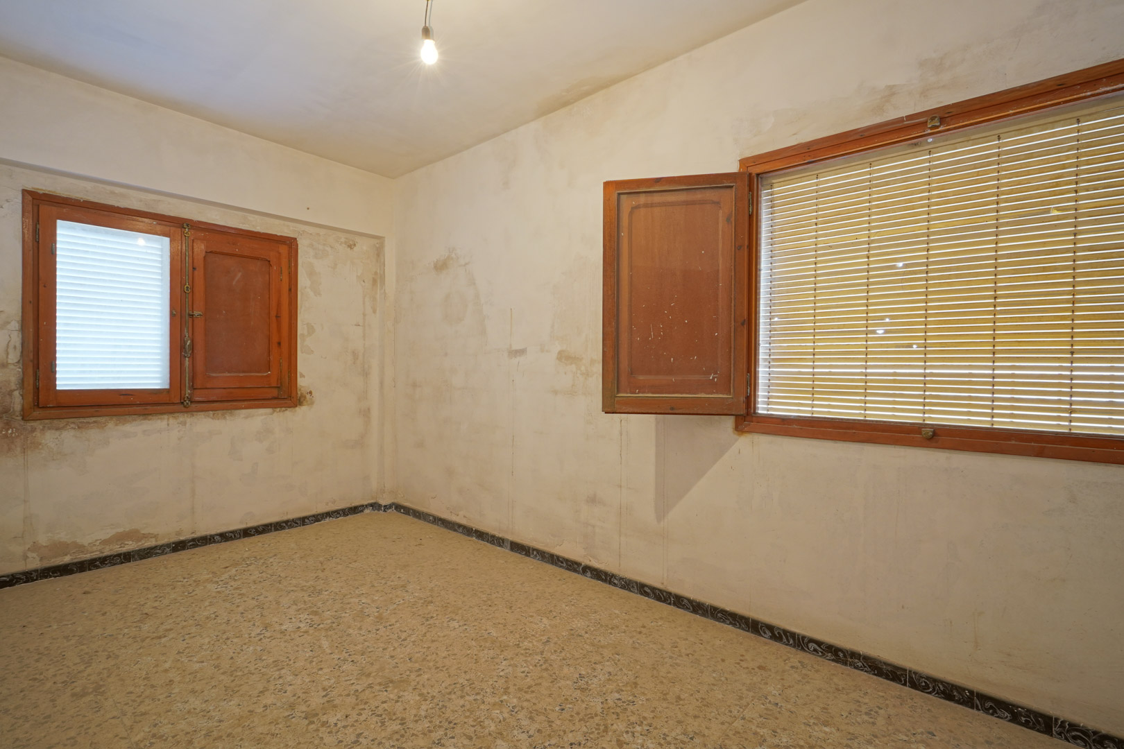 Chambre à rénover avec sol en terrazzo d'origine, fenêtres en bois avec volets et murs offrant un fort potentiel de personnalisation.
