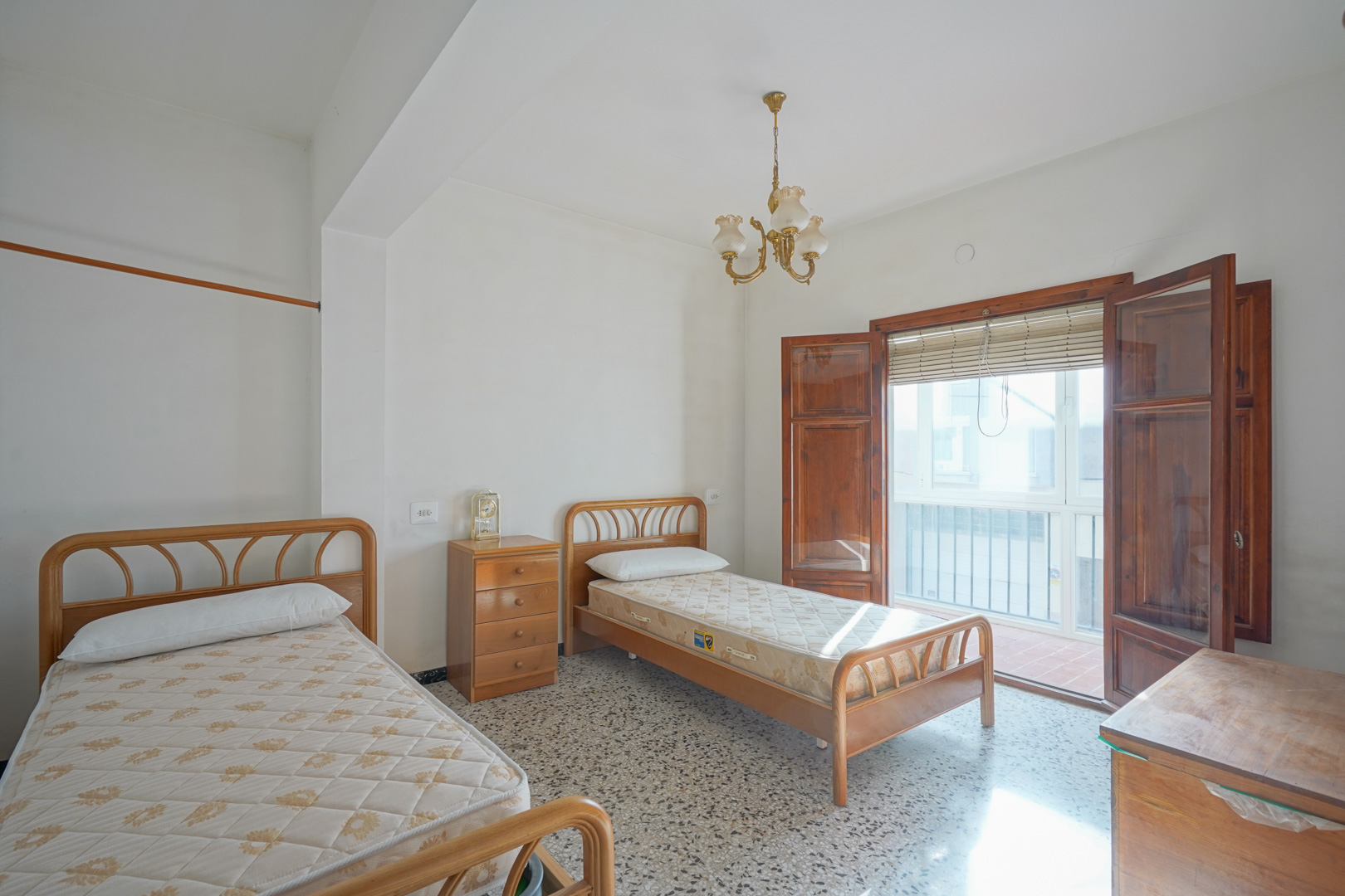 Chambre avec deux lits simples, sol en terrazzo et accès direct à un balcon par des portes-fenêtres en bois massif.
