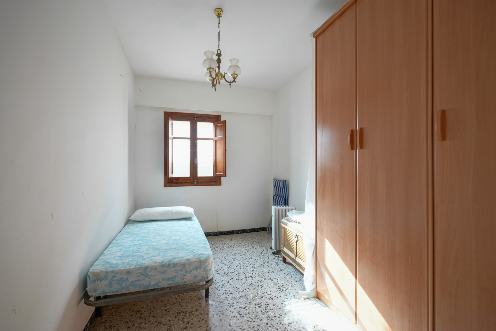 Chambre individuelle avec sol en terrazzo, une grande armoire en bois et une fenêtre en bois naturel apportant une lumière douce.