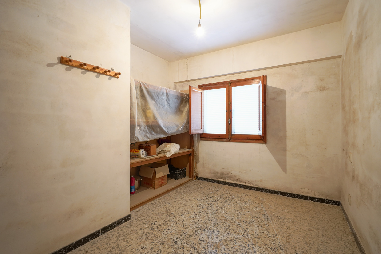 Pièce avec sol en terrazzo, fenêtre en bois à double battant et étagères intégrées, nécessitant une rénovation complète.