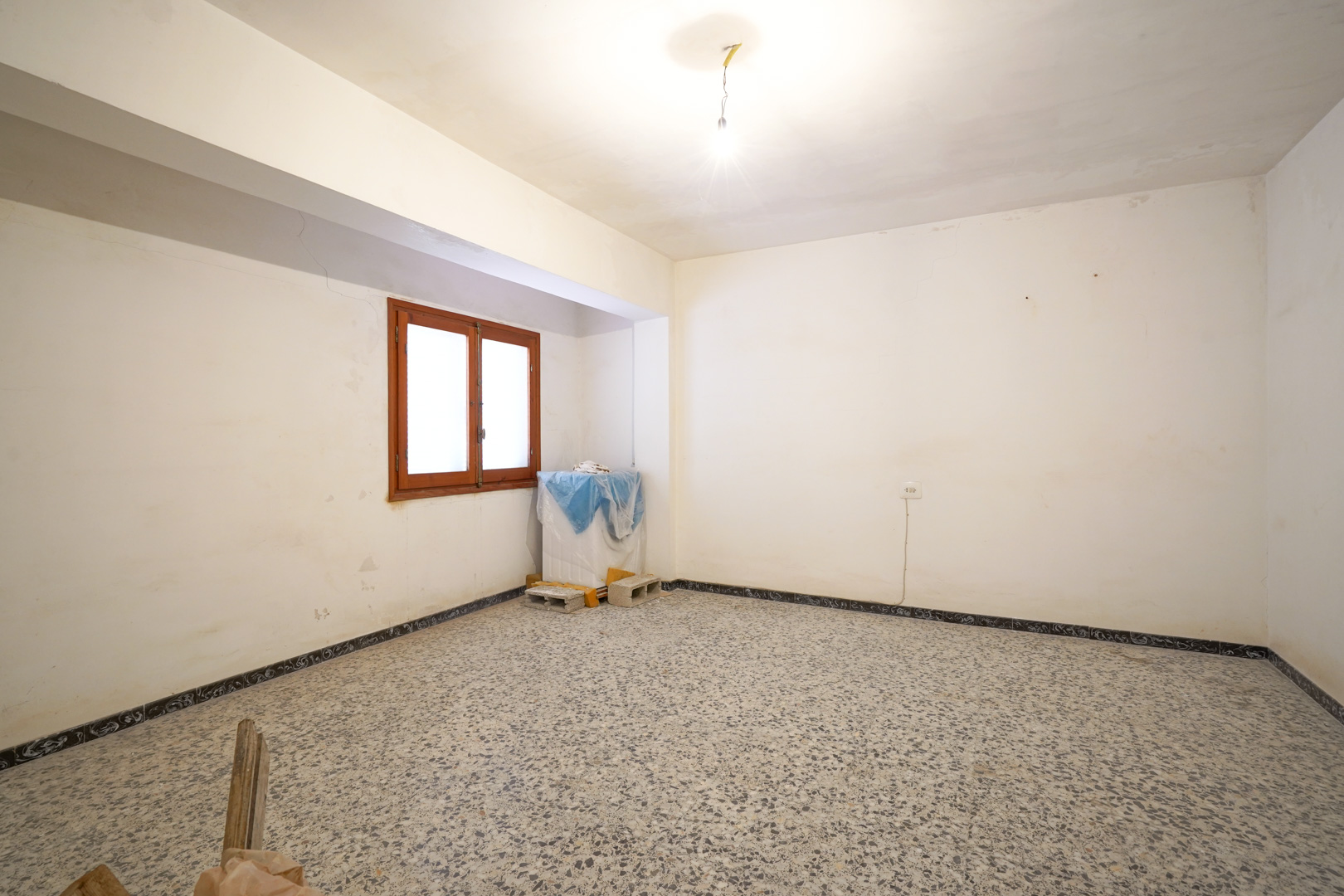 Pièce spacieuse avec sol en terrazzo d'origine, murs blancs à rénover et fenêtre à cadre en bois apportant de la clarté.