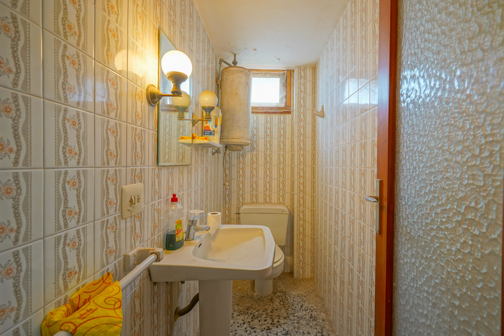 Salle de bain fonctionnelle avec carrelage mural d'époque, lavabo sur pied, chauffe-eau électrique et sol en terrazzo.