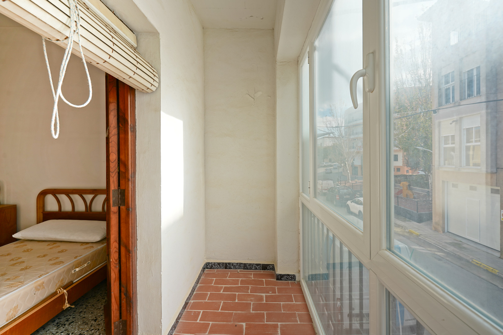 Terrasse vitrée avec carrelage en terre cuite et menuiserie en PVC blanc, offrant une vue sur la rue et un accès direct à une chambre lumineuse.
