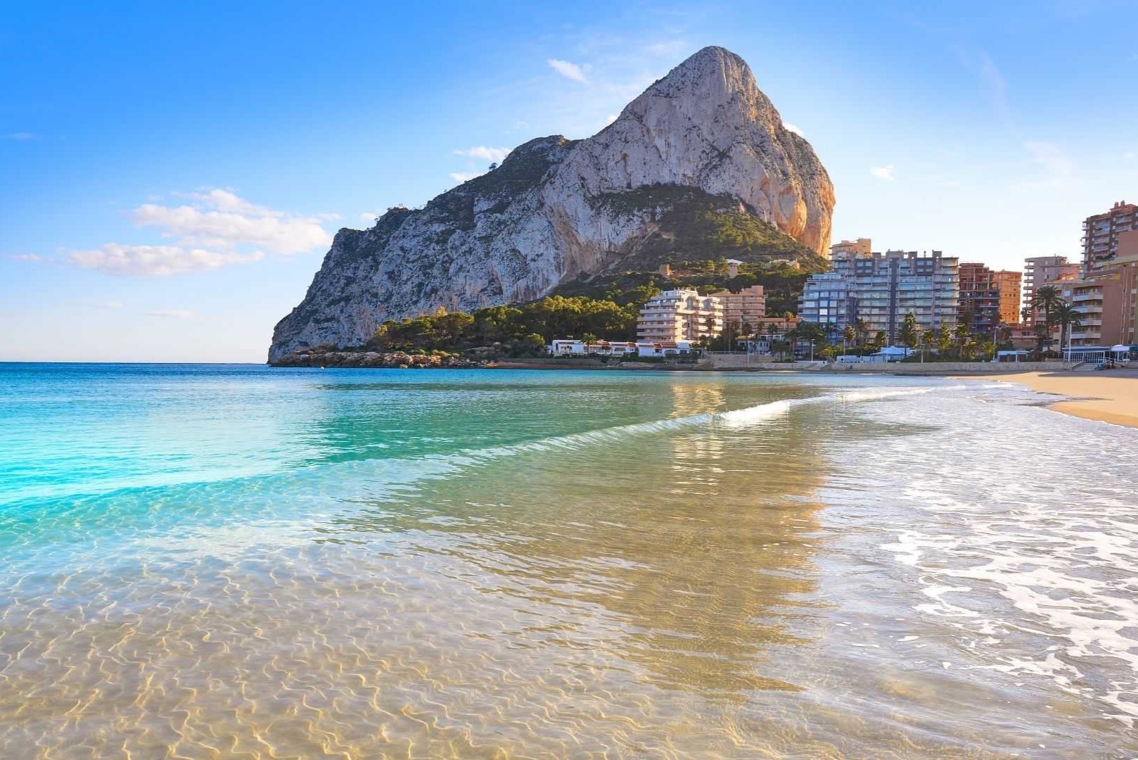 Vue panoramique du Peñón de Ifach depuis le rivage de Calpe, mettant en valeur les eaux turquoise cristallines et le front de mer urbain.