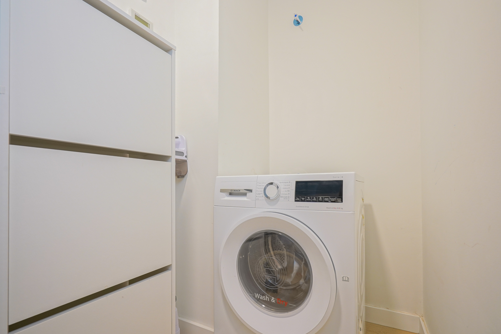 Buanderie fonctionnelle équipée d'un lave-linge séchant Bosch et d'un meuble de rangement blanc, située près du hall d'entrée.
