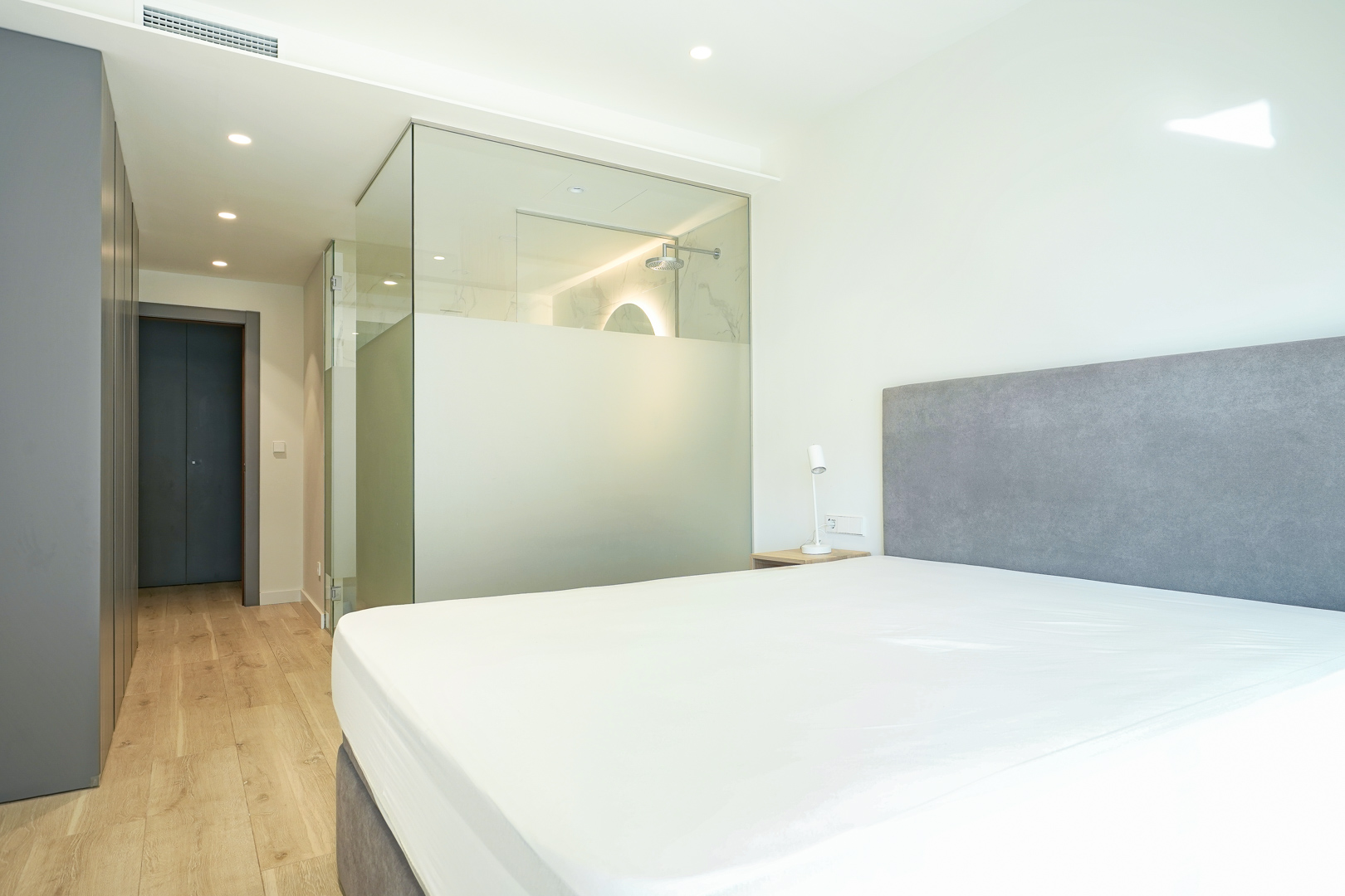 Chambre principale avec salle de bain attenante intégrée par des parois en verre, placards encastrés et parquet clair.