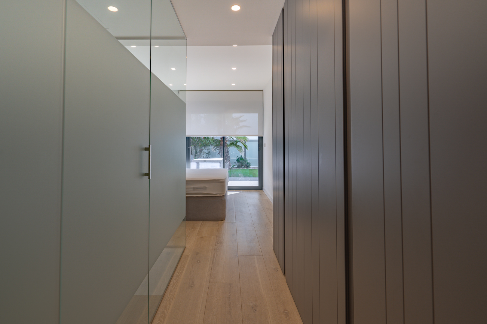 Couloir de la chambre principale avec placards intégrés gris, paroi vitrée et accès visuel au jardin privé par de larges baies vitrées.