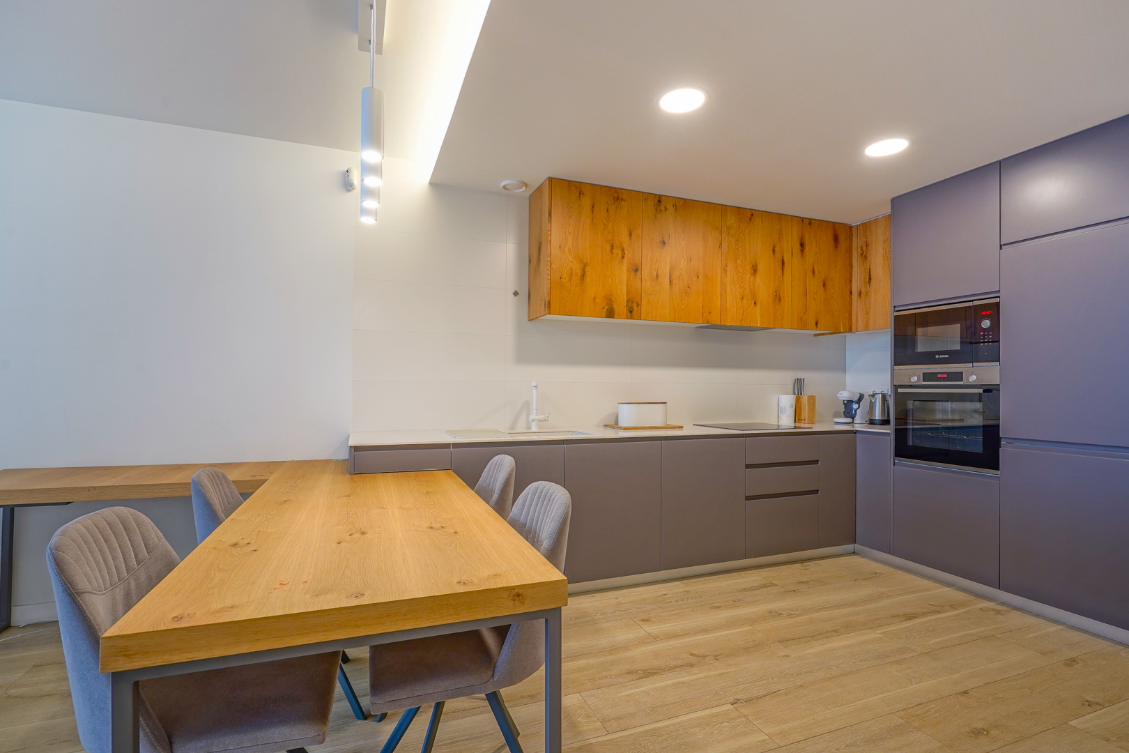 Cuisine moderne à aire ouverte avec placards en bois naturel et gris, électroménagers intégrés et une table à manger en bois assortie avec chaises rembourrées.