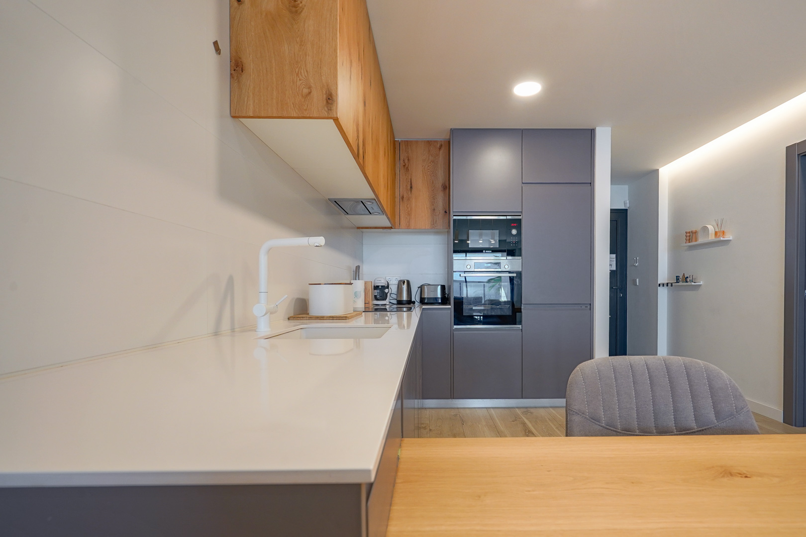 Cuisine moderne avec plan de travail blanc, meubles gris et bois, équipée d'appareils intégrés et d'un robinet blanc minimaliste.