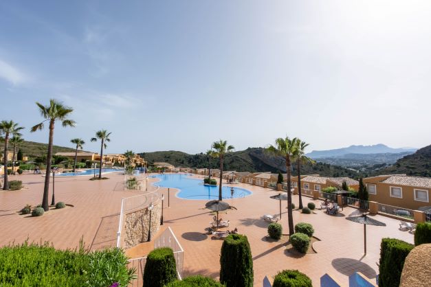 Espace commun de Monte Cala Gardens avec deux grandes piscines, une vaste terrasse ensoleillée bordée de palmiers et une vue dégagée sur les montagnes.
