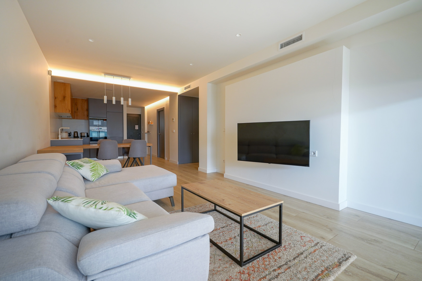 Espace de vie ouvert avec télévision murale, parquet clair et cuisine moderne intégrée dotée d'un éclairage indirect et de finitions en bois.