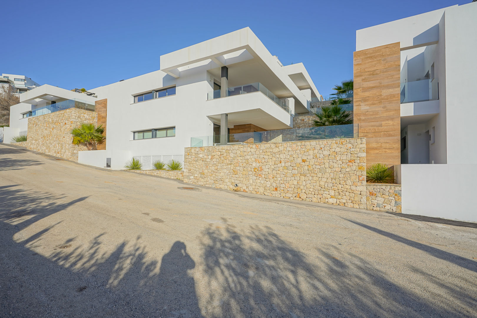 Façade moderne à Monte Cala Gardens avec murs en pierre naturelle, terrasses spacieuses aux garde-corps en verre et finitions élégantes en aspect bois.