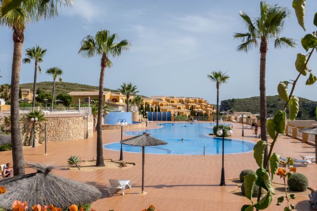 Grande piscine communautaire entourée d'une terrasse en terre cuite, de palmiers et de parasols en bruyère, offrant une vue sur les collines environnantes.