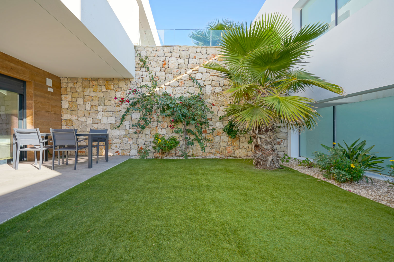Jardin privé avec gazon artificiel, un palmier et un mur en pierre naturelle. Une terrasse couverte abrite un espace dînatoire extérieur aux finitions modernes.