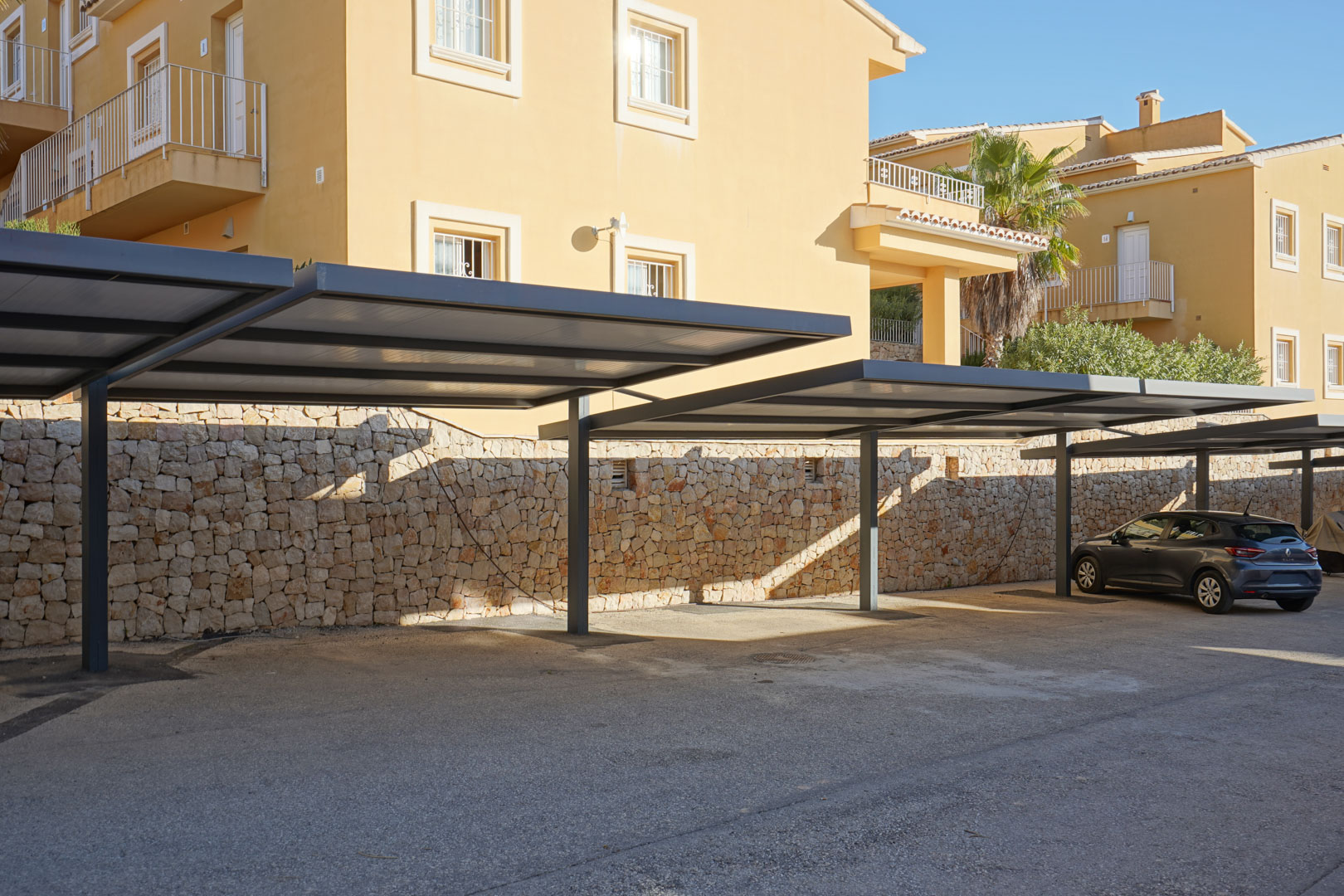 Parking couvert avec structures métalliques modernes et mur en pierre naturelle, situé juste devant l'entrée de la résidence.