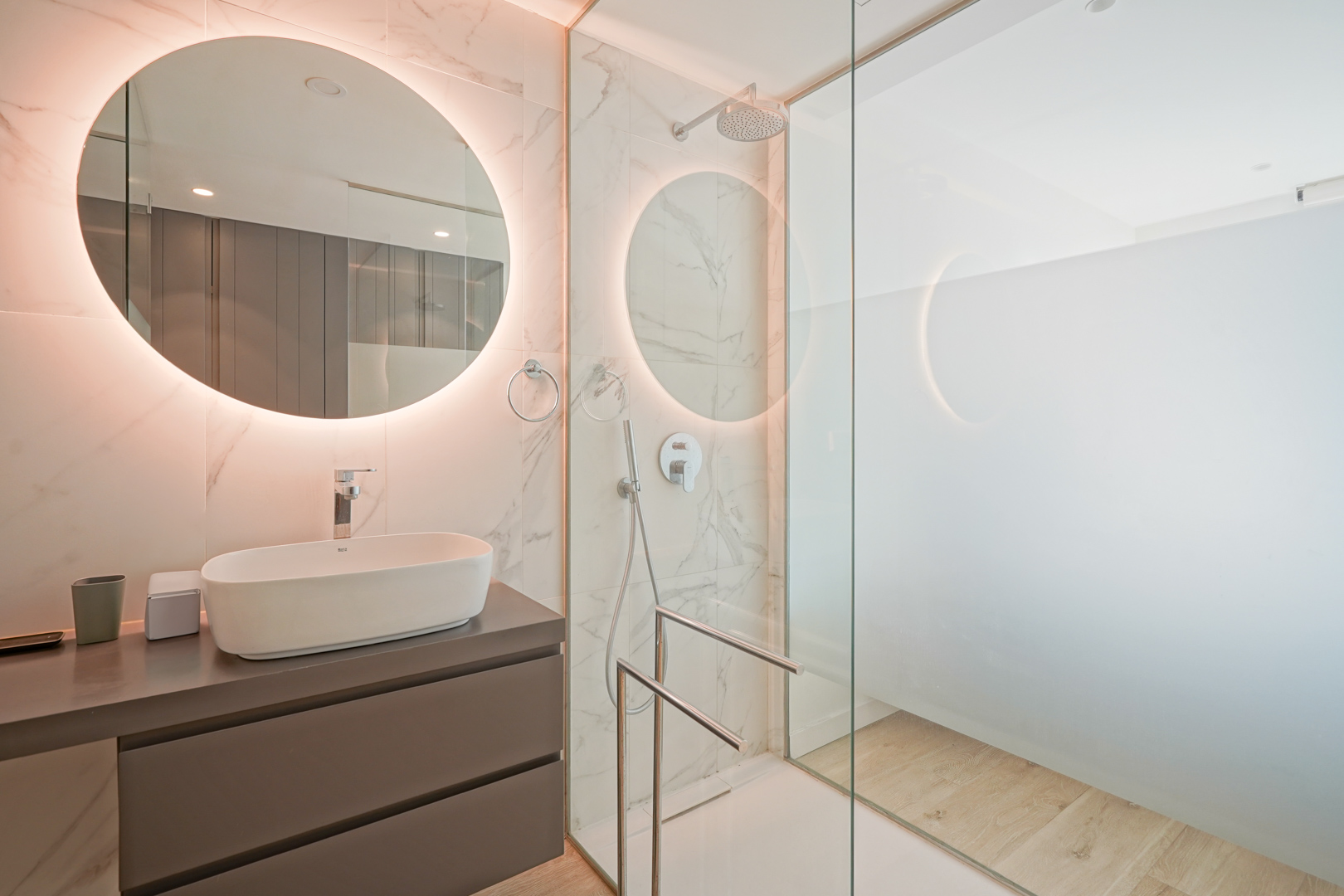 Salle de bain moderne avec vasque à poser, miroir circulaire rétroéclairé et douche à l'italienne avec paroi vitrée et carrelage mural effet marbre.