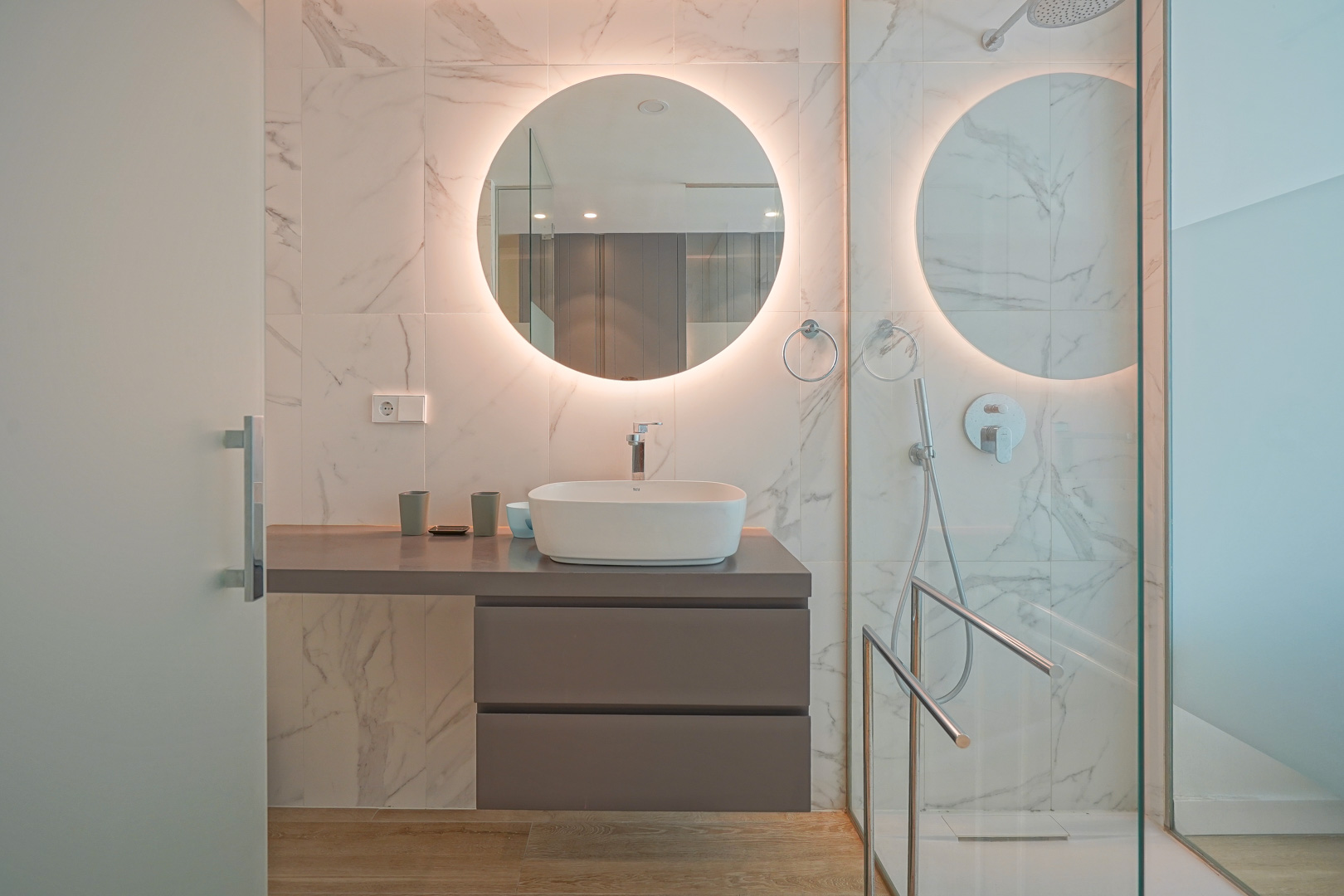 Salle de bain moderne avec vasque à poser, miroir circulaire rétroéclairé et parois en grès cérame effet marbre blanc.