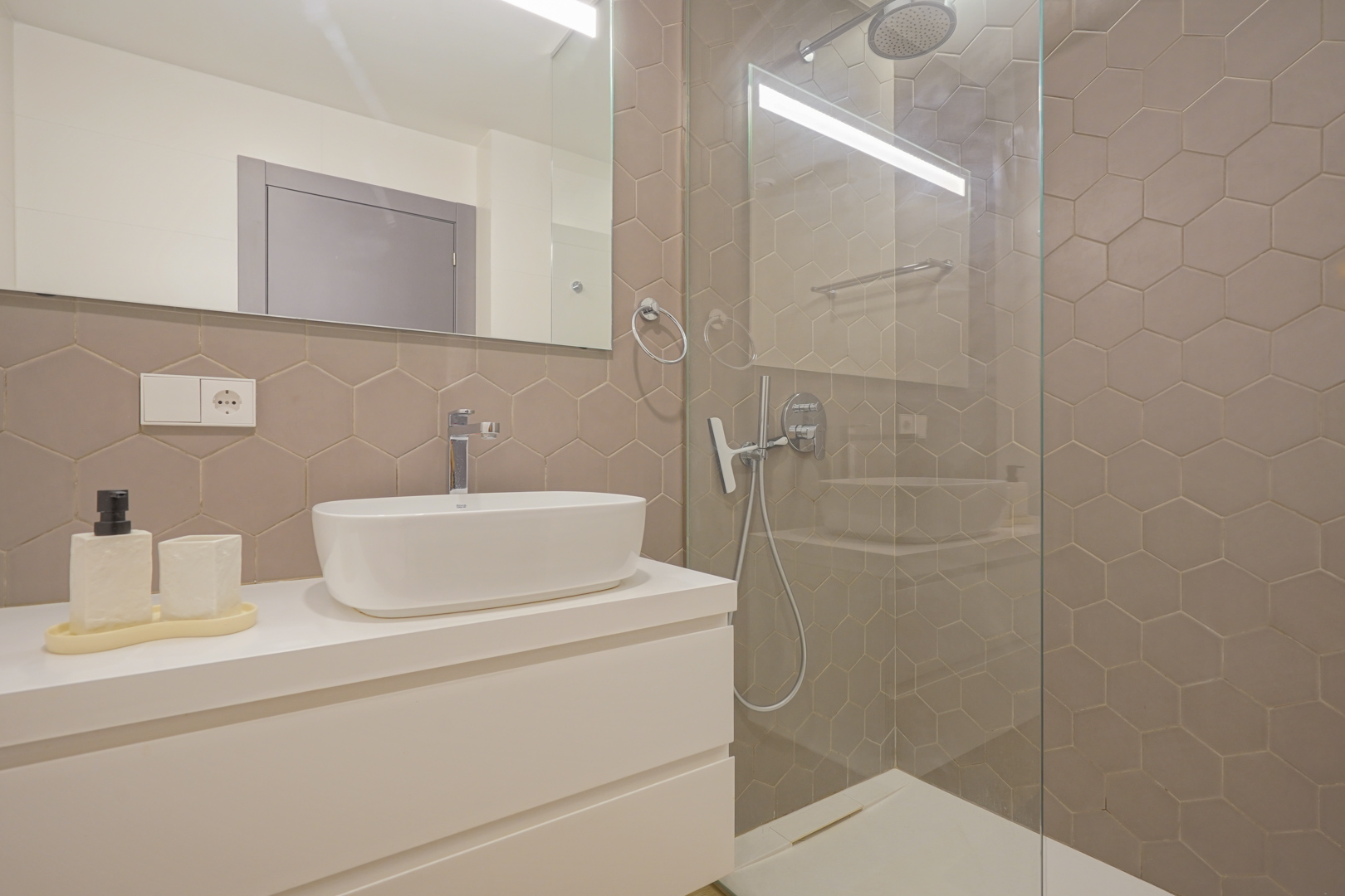 Salle de bains moderne avec carrelage hexagonal taupe, vasque à poser sur meuble blanc et douche à l'italienne avec paroi en verre.