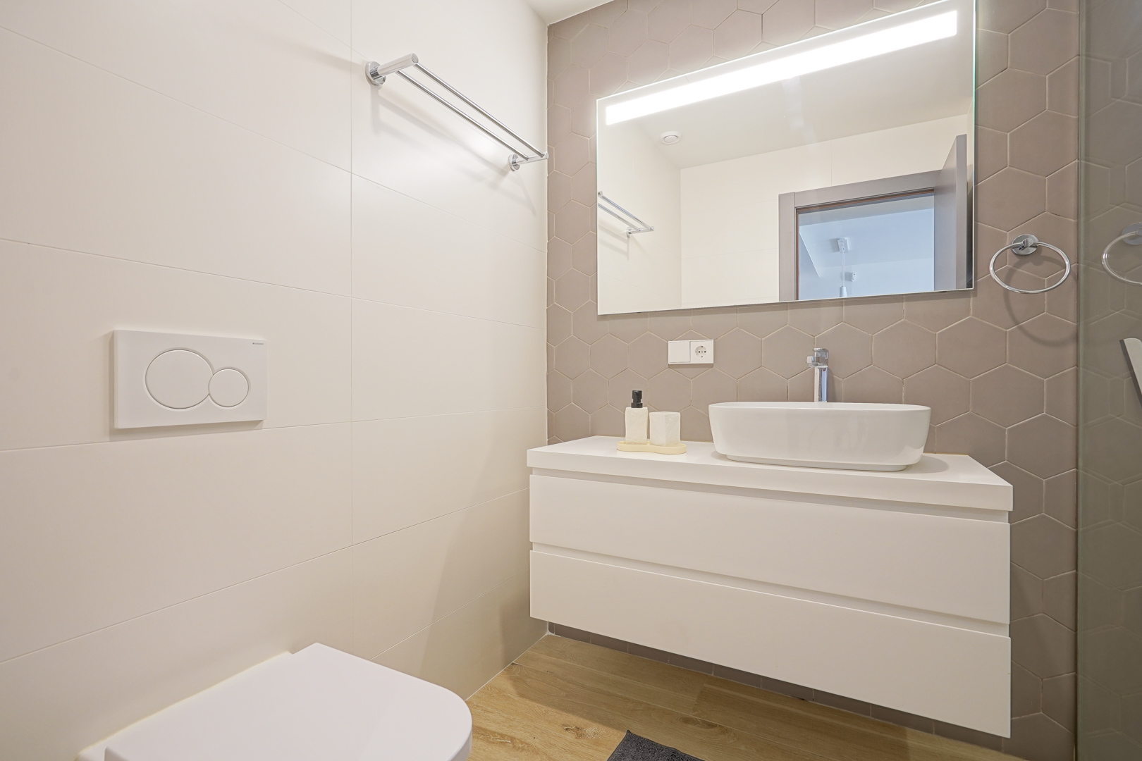 Salle de bains moderne avec crédence en carrelage hexagonal taupe, meuble vasque suspendu blanc et miroir avec éclairage LED intégré. Finitions contemporaines et épurées.