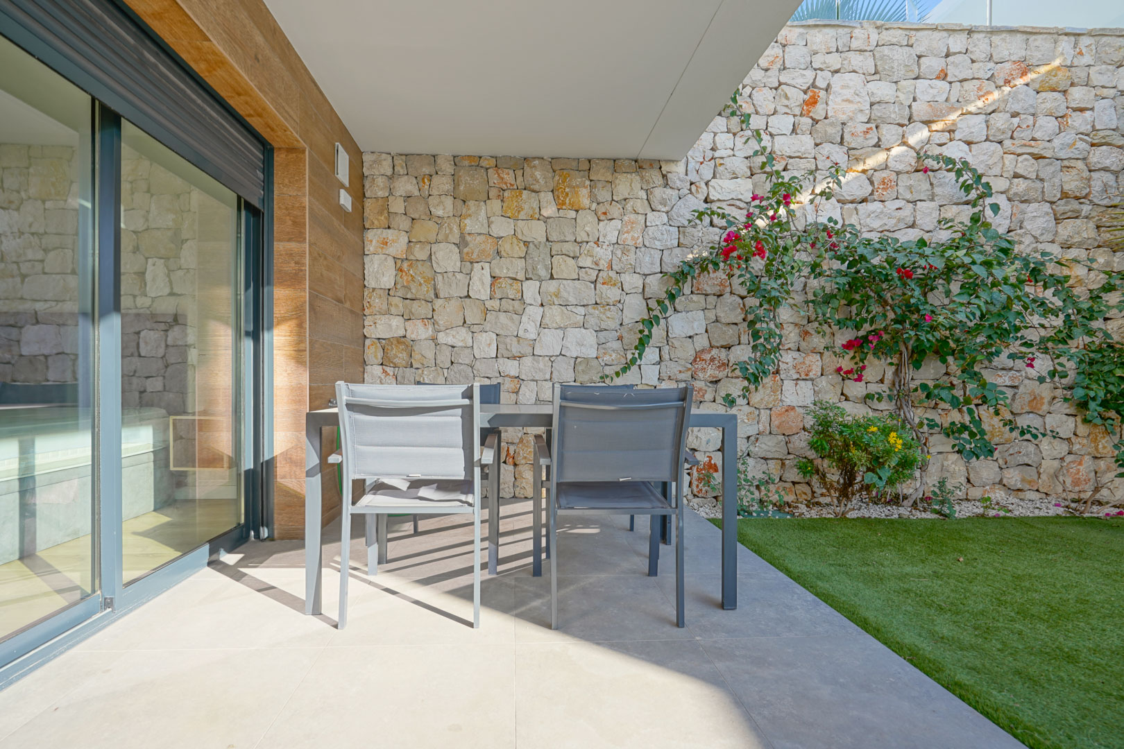 Terrasse couverte avec espace repas extérieur, entourée de murs en pierre naturelle et d'un jardin privé avec gazon synthétique.