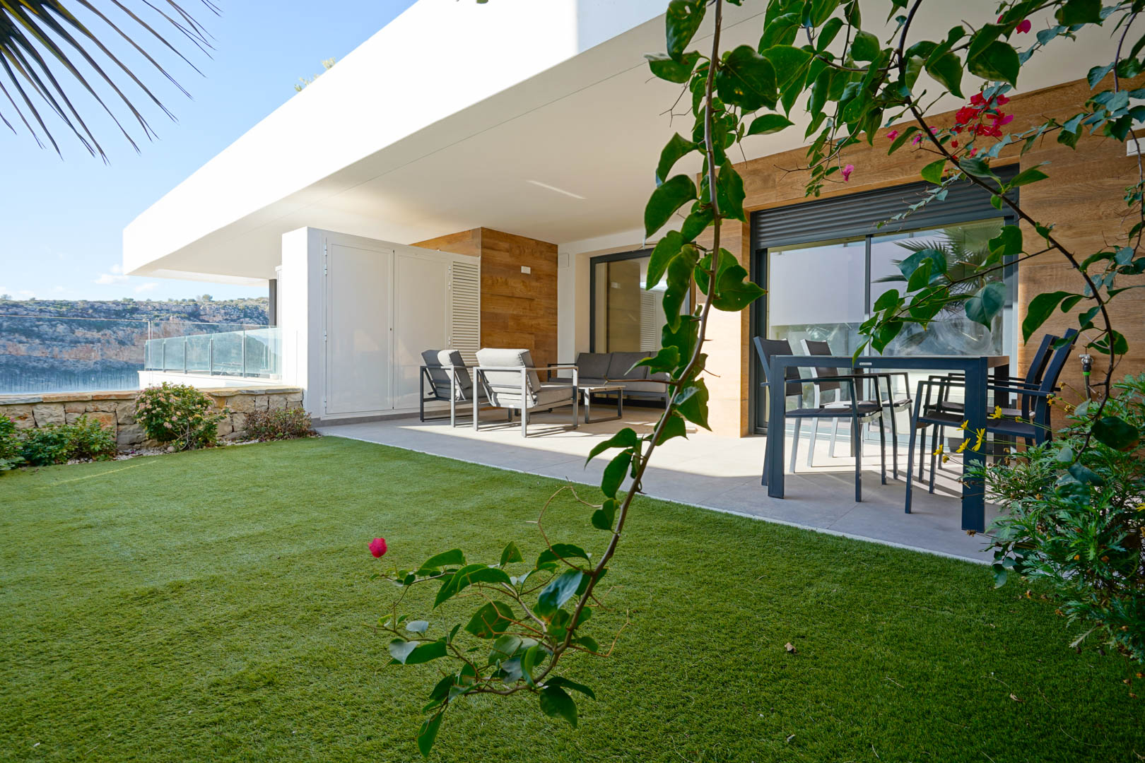 Terrasse couverte avec mobilier de jardin moderne et pelouse privée, bordée d'un mur en pierre naturelle offrant une vue dégagée sur le parc naturel.