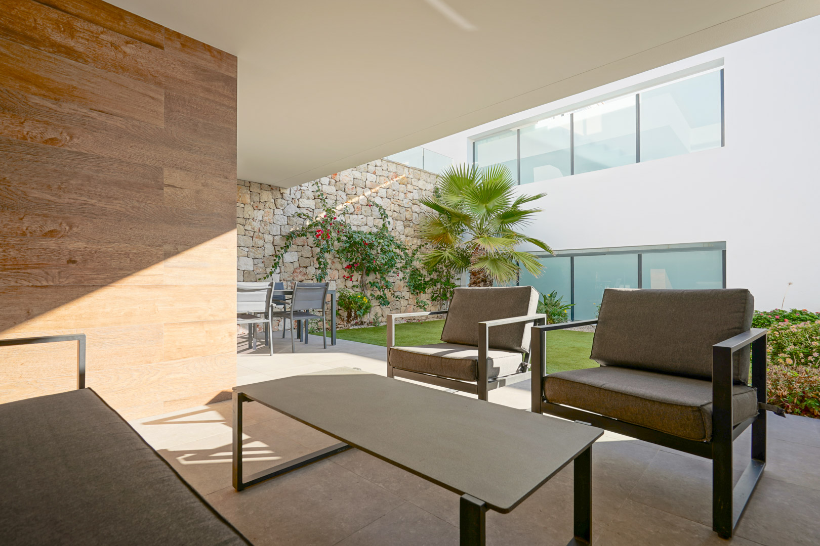 Terrasse couverte avec salon de jardin, coin repas et jardin privé agrémenté d'un mur en pierre naturelle et de palmiers.