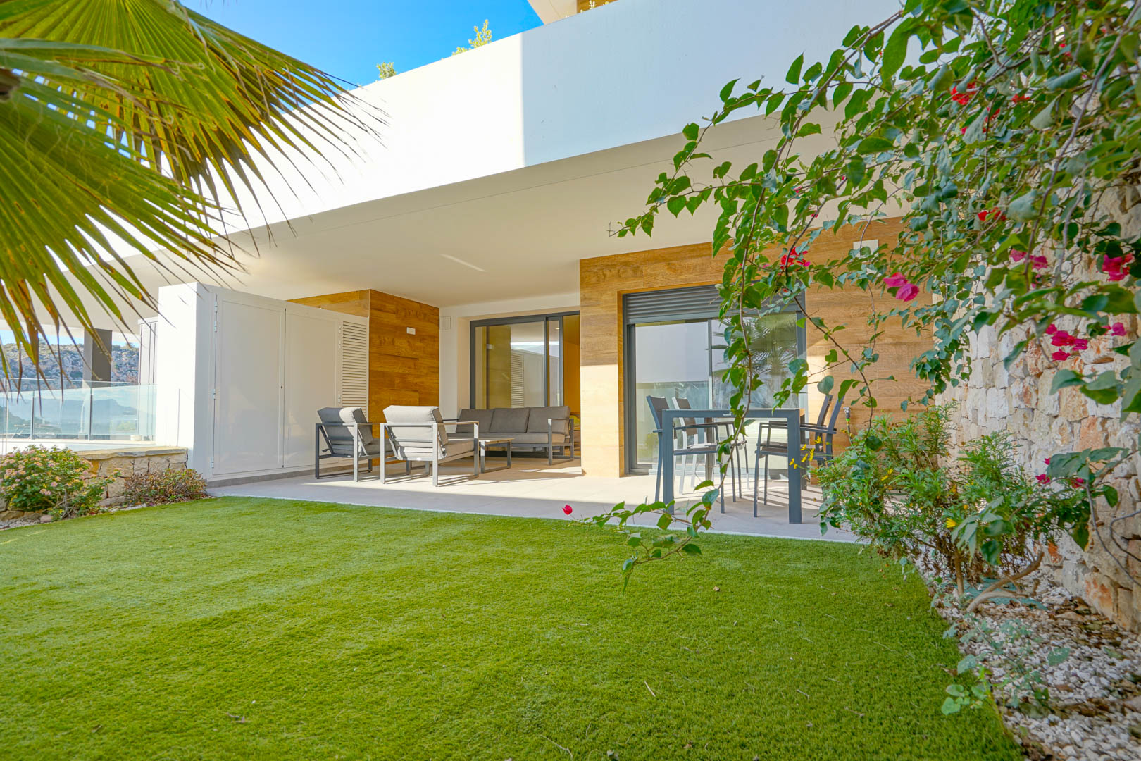 Terrasse couverte avec salon de jardin et coin repas, donnant sur un jardin privé avec pelouse artificielle et mur en pierre naturelle.