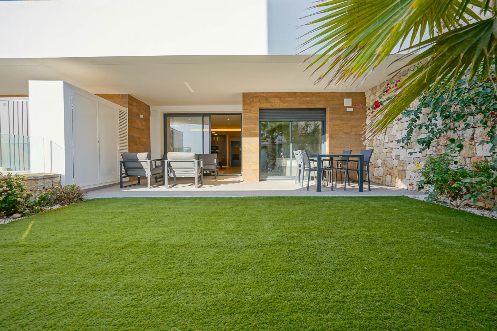 Terrasse couverte avec salon de jardin et espace repas, s'ouvrant sur un jardin privé avec pelouse et mur de clôture en pierre naturelle.