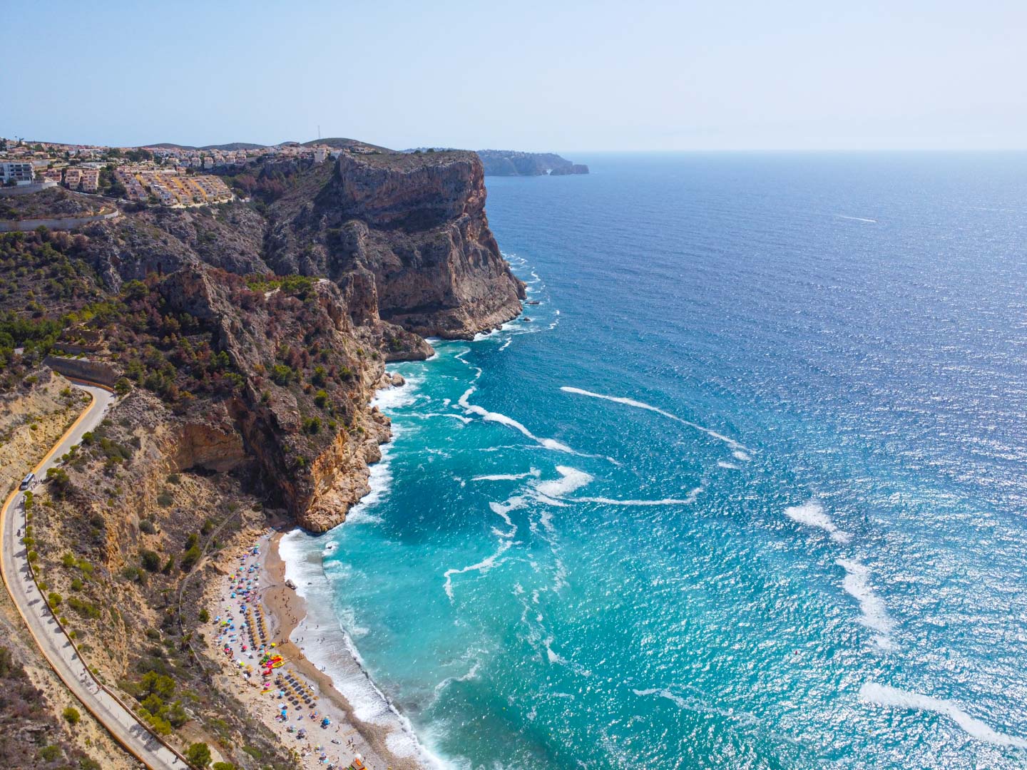 Vue aérienne de la Cala del Moraig à Cumbre del Sol, montrant les falaises escarpées, les eaux turquoise et la route sinueuse menant à la plage.