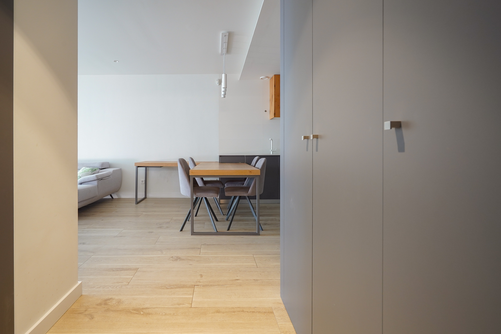 Vue de l'entrée avec placards gris intégrés, parquet clair et espace repas ouvert comprenant une table en bois et des chaises contemporaines.
